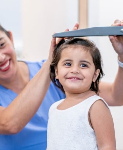 A sweet little girl of Indian decent, stands up tall as her doctor measures her height.  She has a light summer dress on and is smiling at her female doctor.  The doctor is wearing blue scrubs and is holding the measuring tool gently above the little girls head as she takes not of the number.